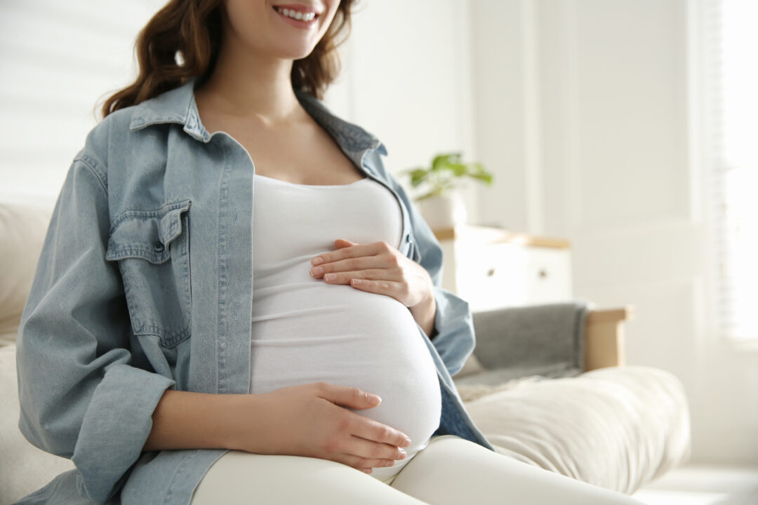 Schwangere Frau in weißem Shirt sitzt auf dem Sofa und umfasst ihren Bauch